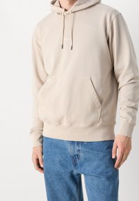 Beige hoodie i mjukt tyg, med en framficka, ribbad mudd vid ärmsluten och justerbara dragsnören i huvan. Kombinerad med blå jeans.