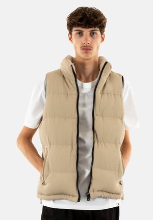 Gilet matelassé beige avec texture à côtes verticales, fermeture éclair sur le devant et col montant. Dispose de poches latérales et d'un logo discret sur la poitrine.