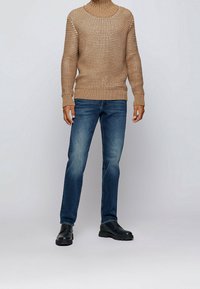 Béžový pletený svetr s vysokým límcem, v kombinaci s modrými džíny z denimu. Model nosí černé kožené boty.