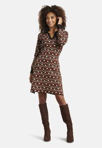 Langärmliges, V-Ausschnitt-Kleid in Braun mit einem geometrischen Muster in Creme, Schwarz und Rot. Getragen mit knielangen, braunen Wildlederstiefeln.