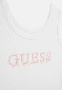 White ribbed tank top featuring pink embroidered "GUESS" logo on the front. Round neckline and thin straps. Smooth texture.