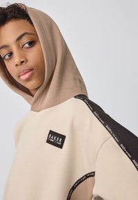 Beige og brun hættetrøje med et rektangulært logopatch, kontrasterende teksturstof og mønstrede detaljer på ærmerne.