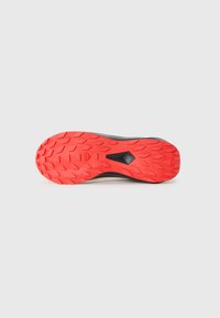 Semelle de chaussure de sport noire avec un motif de traction en caoutchouc rouge proéminent, présentant une texture en losanges et une découpe centrale pour la flexibilité.