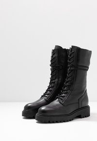 Bottes de combat en cuir noir avec une finition texturée, devant lacé et semelle robuste. Caractérisées par une tige haute et des détails cousus sur les côtés.