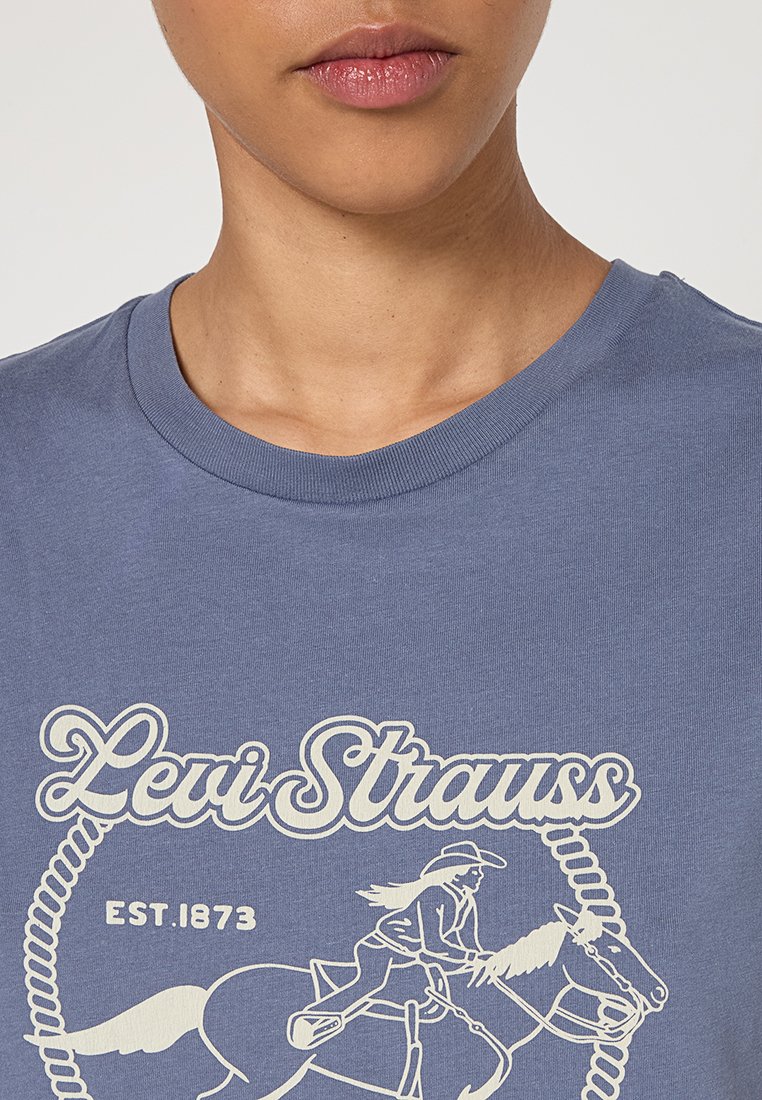Person, der ein blaues Levi Strauss T-Shirt mit weißem Logo trägt, das einen Reiter auf einem Pferd und den Text "EST. 1873" zeigt.