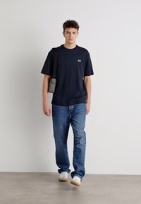 Tricou negru cu mânecă scurtă, având un logo mic, asortat cu blugi albaștri deschis și adidași albi. Accesoriile includ o geantă mică de umăr.