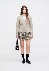 Jovem mulher vestindo um cardigã de malha bege, saia xadrez, meias e sapatos pretos, segurando uma bolsa preta, de pé contra um fundo liso.