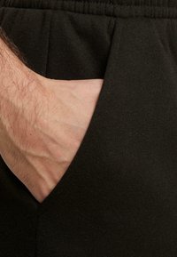 Des pantalons de jogging en tissu noir présentent une poche latérale avec une texture lisse, une taille élastique et un gros plan d'une main partiellement insérée dans la poche.