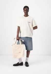Kreemipiguline t-särk koos "Originals" tikandiga, sinised denim-lühikesed püksid, beež kangast kott punase "Levi's" logoga, mustad loafersid, valged sokid.