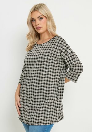 Frau mit blondem Haar, die eine schwarz-weiß karierte lange Tunika mit seitlichen Knöpfen und blaue Jeans trägt, vor einem einfarbigen Hintergrund stehend.