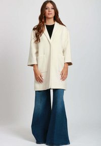 Cappotto in misto lana color crema con colletto a scialle, maniche a tre quarti e due tasche frontali, abbinato a jeans svasati blu.