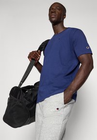 Camiseta de algodão azul com decote redondo e logótipo, combinada com calças de moletão cinza claro. Uma mala de viagem preta é segurada pela alça de ombro.
