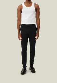 Schwarze, maßgeschneiderte Hosen mit glatter Textur, kombiniert mit einem weißen ärmellosen Tanktop. Das Outfit wird durch schwarze Schnürschuhe ergänzt.