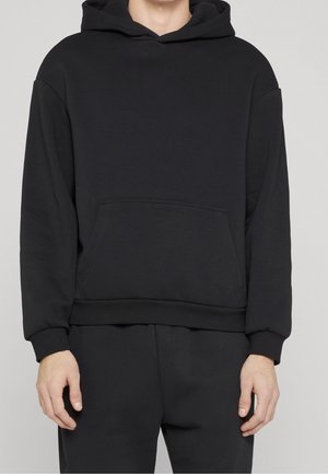 Hombre vistiendo una sudadera negra lisa con capucha, con un bolsillo frontal estilo canguro, y pantalones negros a juego, sobre un fondo gris claro.