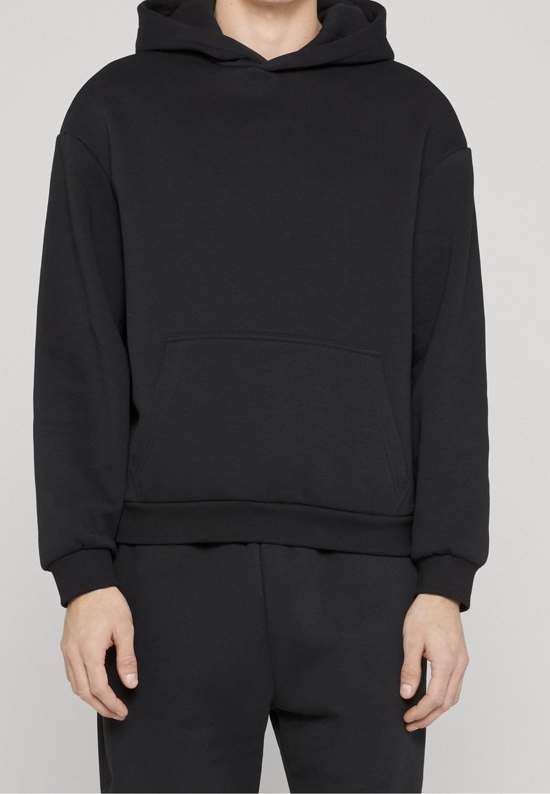 Homme portant un sweat à capuche noir uni avec une poche kangourou avant et un pantalon noir assorti, sur un fond gris clair.