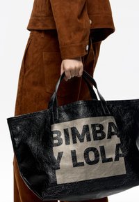 Didelis juodas tote krepšys su blizgia tekstūra, turintis išsiskiriantį smėlio spalvos skyrių su tekstu "BIMBA Y LOLA" ryškiai juodomis raidėmis.