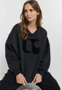 Svart oversized sweatshirt med en stor svart logotyp framtill. Tyget ser mjukt ut, med sänkta axlar och långa ärmar.