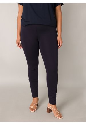 Vrouw die zwarte nauwsluitende broek en beige sandalen met hak en bandjes draagt, staand tegen een lichte achtergrond.