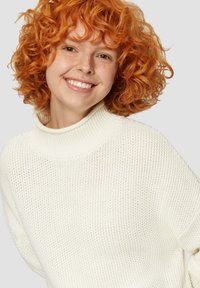 Biały, dziany sweter z wysokim kołnierzem i luźnym krojem. Posiada teksturowany wzór i ściągacz u dekoltu, noszony przez osobę z kręconymi, pomarańczowymi włosami.