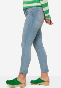 Jeans ajustados de color azul claro con detalles desgastados, puños arrollados, cinco bolsillos, combinados con zuecos de ante verde y un suéter a rayas verdes.