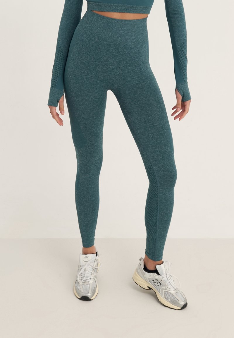 Světlé modré legíny s vysokým pasem a bezešvou konstrukcí, vyrobené z texturované látky, v kombinaci se šedými sportovními botami.