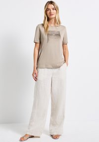 Vaaleanbeige t-paita lyhyillä hihoilla, jossa on tummemmista sävyistä koostuva painatus, yhdistettynä vaaleisiin, löyhiin pellavahousuihin ja remmisandaaleihin.