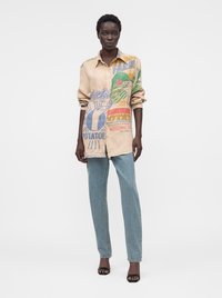 Mujer de pie con camisa beige de gran tamaño con botones y estampados de granja y patatas, vaqueros azul claro y tacones negros con punta abierta.