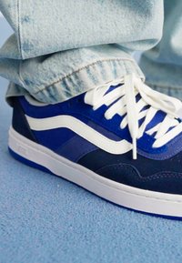 Blue suede sneaker with white accents, featuring a low top design and white laces, set against a light blue textured surface.
