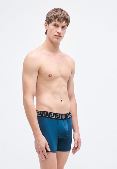 Τυρκουάζ κοντά εσώρουχα τύπου μποξεράκια με μαύρη μέση που έχει γεωμετρικό μοτίβο. Το ύφασμα φαίνεται λείο με ελαφριά γυαλάδα.