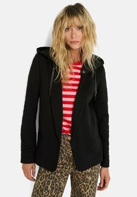 Blazer negro con capucha, con una tela texturizada y cordones brillantes. Lleva puesto una camiseta a rayas rojas y pantalones de estampado de leopardo.