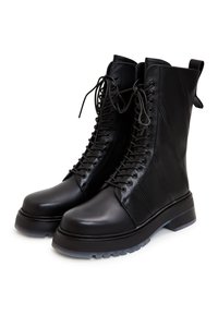 Botas altas de cuero negro hasta la rodilla con caña alta, punta redonda, suela gruesa y cordones con ojetes. Presentan paneles laterales texturizados y una lengüeta para facilitar el calzado.