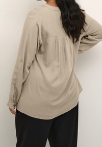 Beżowa koszula z długim rękawem, z plisowanym tyłem i mankietami na guziki, wykonana z lekkiego materiału. W połączeniu z czarnymi spodniami.