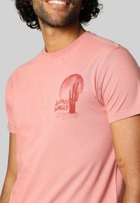 T-shirt en coton rose avec un graphisme de coucher de soleil et de cactus, accompagné du texte "SAFARI SUNSET" en rouge. Manches courtes, design col rond.