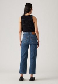 Kvinna som står med ryggen mot, iklädd en svart ärmlös stickad topp, högmidjade blå raka jeans och svarta loafers på en grå bakgrund.