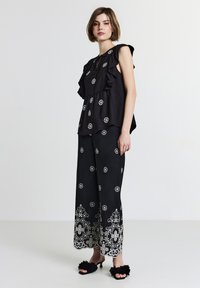 Donna che indossa un top nero senza maniche con volant e pantaloni larghi abbinati, decorati con ricami floreali bianchi, indossati con sandali con tacco florali neri.