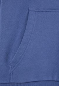 Tessuto di felpa blu con tasca frontale. Texture liscia, orlo a coste. Nessun motivo o dettaglio visibile. Focalizzazione su materiali e dettagli di costruzione.
