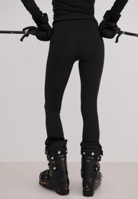 Sorte tettsittende leggings kombinert med svarte skistøvler med sølvdetaljer og et strømlinjeformet design, holdt med skistaver, mot en nøytral bakgrunn.