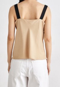 Beige ärmlös topp med svarta band, med en avslappnad passform och slät textur. Bärs med vita högmidjade byxor.