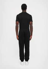 Camiseta de algodón negra y pantalones jogging negros con cinturilla elástica, con puños en los tobillos. Zapatillas blancas completan el conjunto. Vista trasera.