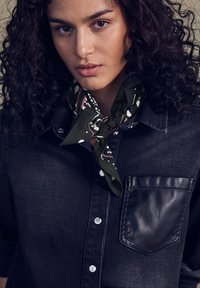 Camicia in denim scuro con una tasca in pelle nera, caratterizzata da bottoni argentati, abbinata a un foulard a motivi verde scuro annodato al collo.