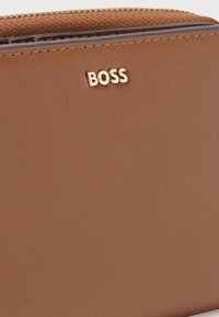 Κοντινή λήψη καφέ δερμάτινου πορτοφολιού με χρυσό λογότυπο "BOSS" και ορατό φερμουάρ στην υφασμάτινη επάνω άκρη.