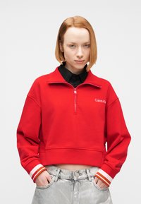 Νεαρός με κοντά κόκκινα μαλλιά, φορώντας κόκκινο φούτερ Calvin Klein με μισή ζώνη και ασημί παντελόνι, με τα χέρια στις τσέπες.