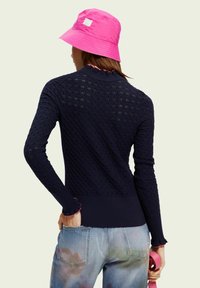 Granatowy sweter dziergany z teksturowanym wzorem diamentowym, ściągaczami na mankietach i dole. Połączony z jasnoniebieskimi dżinsami z kolorowymi akcentami. Różowy kapelusz typu bucket.