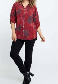 Femme portant un chemisier rouge à fleurs boutonné avec manches retroussées, pantalon noir et bottines noires à talons, sur fond blanc uni.