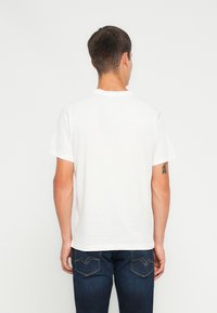 Camiseta de algodón blanca con cuello redondo y mangas cortas, con un diseño liso. Combinada con jeans de color azul oscuro y un tatuaje visible en el brazo.