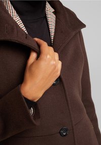 Abrigo de lana marrón con un cuello de patrón de espiga. Presenta un cuello con muescas y botones, con un primer plano de una mano ajustando el cuello.