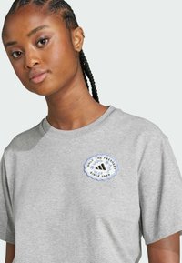 Grå bomullströja med rund broderad patch med text och logotyp i blått och svart, korta ärmar och klassisk passform.