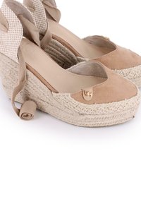 Zapatos de cuña de esparto de gamuza beige con una base de yute trenzado, que cuentan con una punta redondeada, ataduras en el tobillo y un pequeño logotipo metálico como acento.