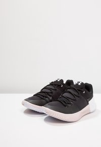 Un paio di sneaker da atletica Under Armour bianche e nere con suole imbottite e lacci, posizionate su una superficie bianca semplice.