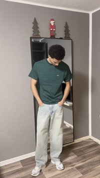 Grün und marineblau gestreiftes T-Shirt, lockere Passform; hellblaue, leicht ausgewaschene Jeans mit geradem Bein; weiße und graue Sneaker; großer Spiegel im Hintergrund.